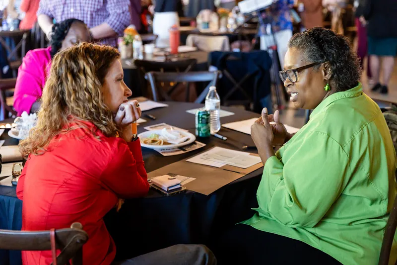 Two attendees in conversation across a round table in the event hall, one leaning in to listen.
