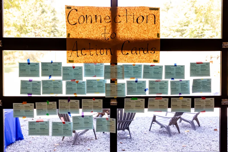 A hand-lettered “Connection to Action Cards” sign above rows of filled-in cards taped to a window, with the gravel patio and Adirondack chairs outside.