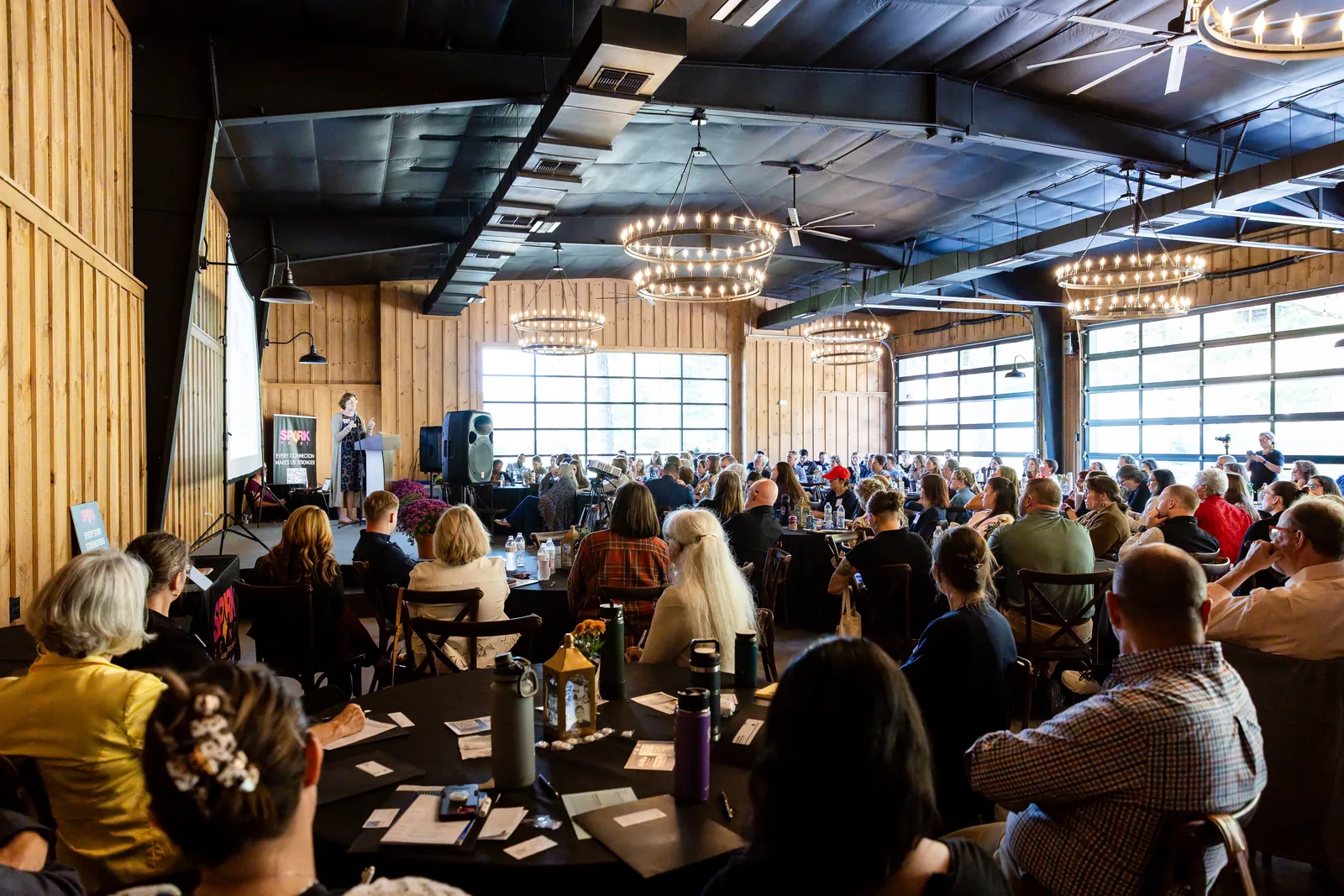 Seen from the back of the hall, a full room at round tables faces a speaker at the podium beneath wagon-wheel chandeliers.