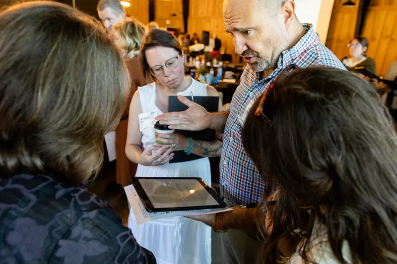 A small group gathers around a tablet as one participant points to the screen while working through the Rural Health Field Simulator.