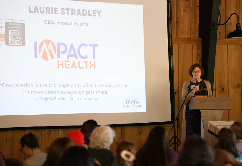 Laurie Stradley, CEO of Impact Health, speaks from the podium beside a slide quoting Virginia Burden—“Cooperation is the thorough conviction that nobody can get there unless everybody gets there”—as a full room listens.