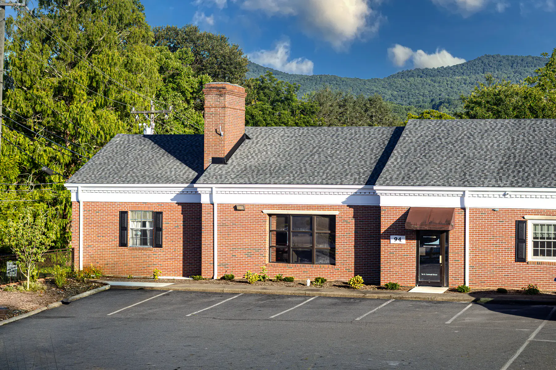 The building at 94 S. Caldwell Street today: a one-story brick office with a chimney, an empty parking lot, and the Blue Ridge behind it.